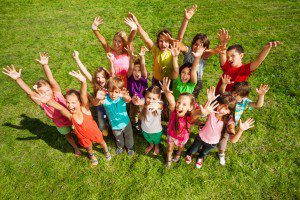 Large group of happy kids, boys and girls, with lifted hands about 10 years old standing on the green grass top view