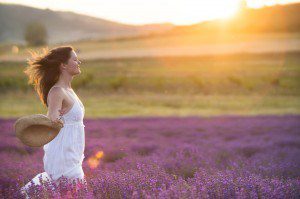 Beautiful young healthy woman with a white dress running joyfully through a lavender field holding a straw hat under the rays of the setting sun.
